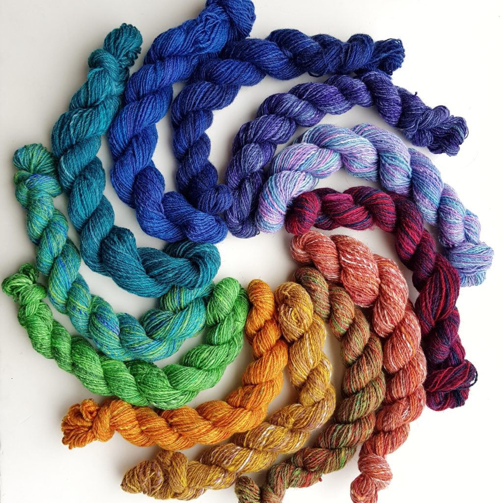 Twelve mini skeins of yarn, arranged in a circle, radiating out. They are ordered by colour, forming a rainbow.