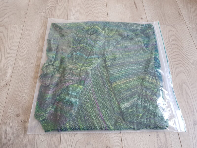 A green hand knit shawl inside a zip loc bag.